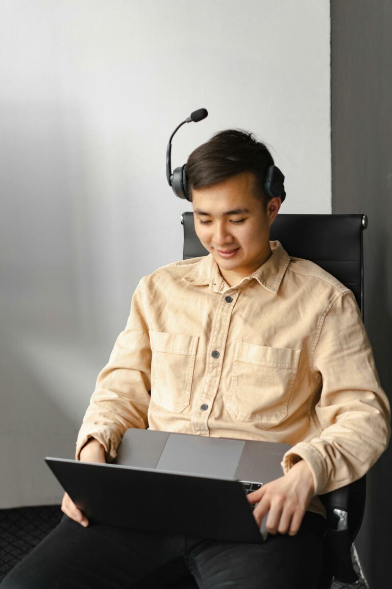 Asian man wearing headset, working on a laptop in an office environment, communicating online.